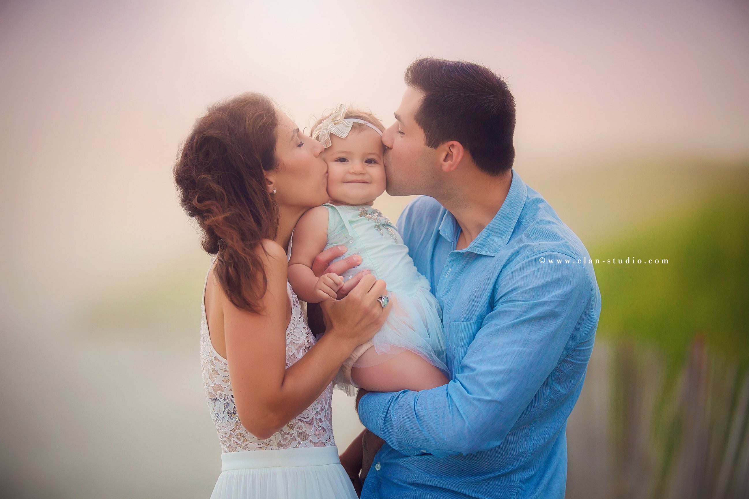 mother and father kissing baby girl at sunset