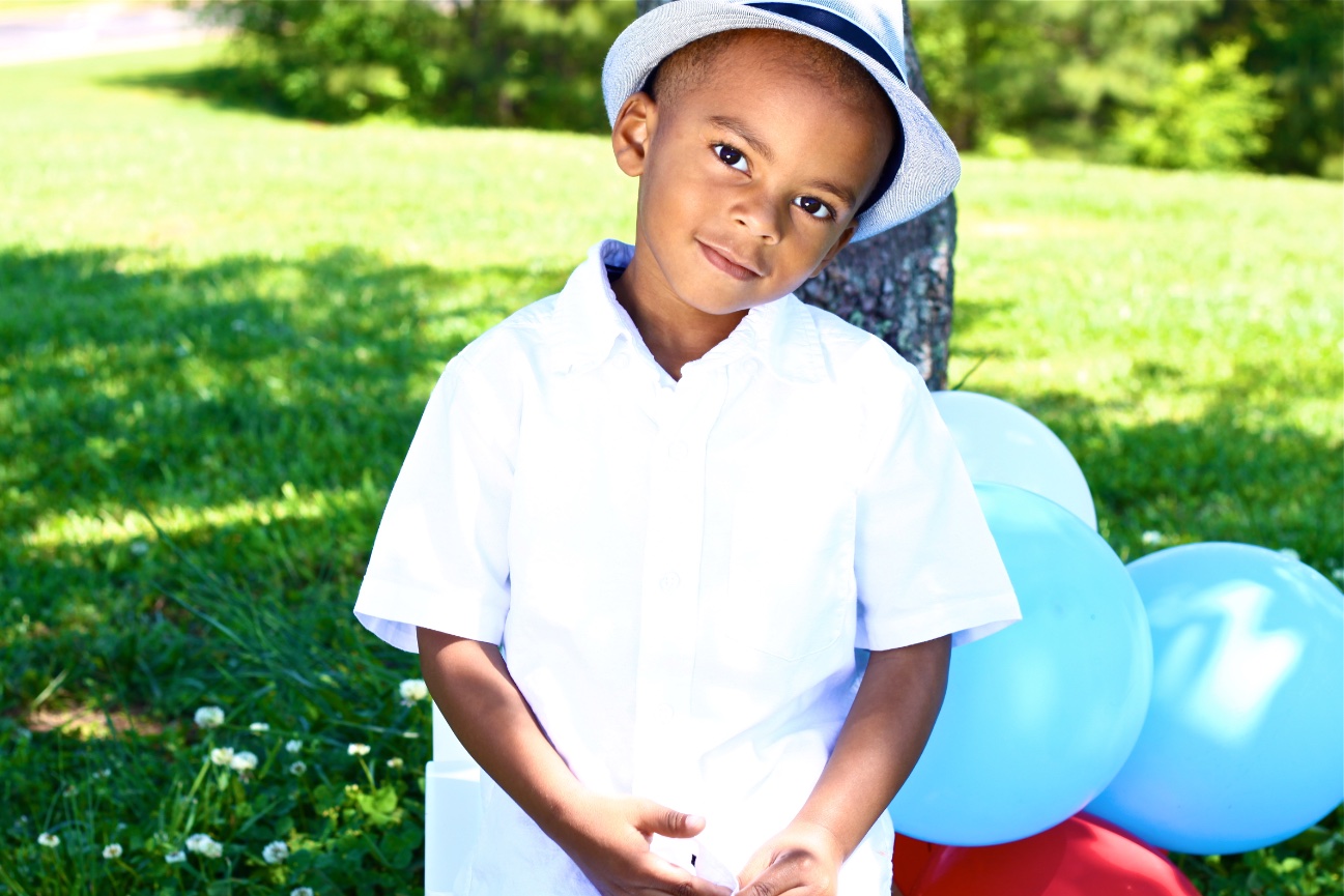 Young boy standing outdoors beside blue balloons in a sunny park.