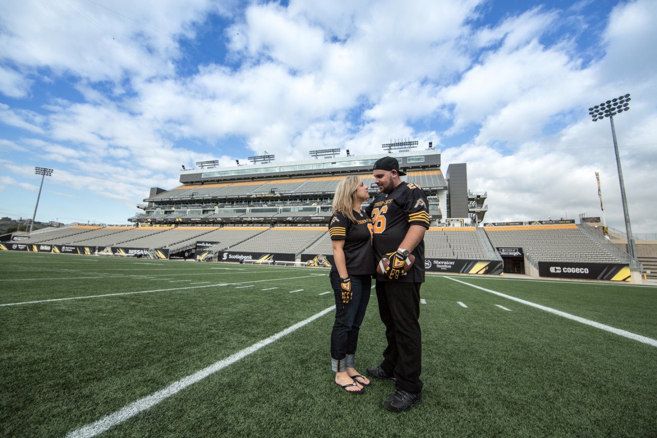 A couple stands on a football field with an empty stadium in the background under a blue sky. Tim Horton's Field