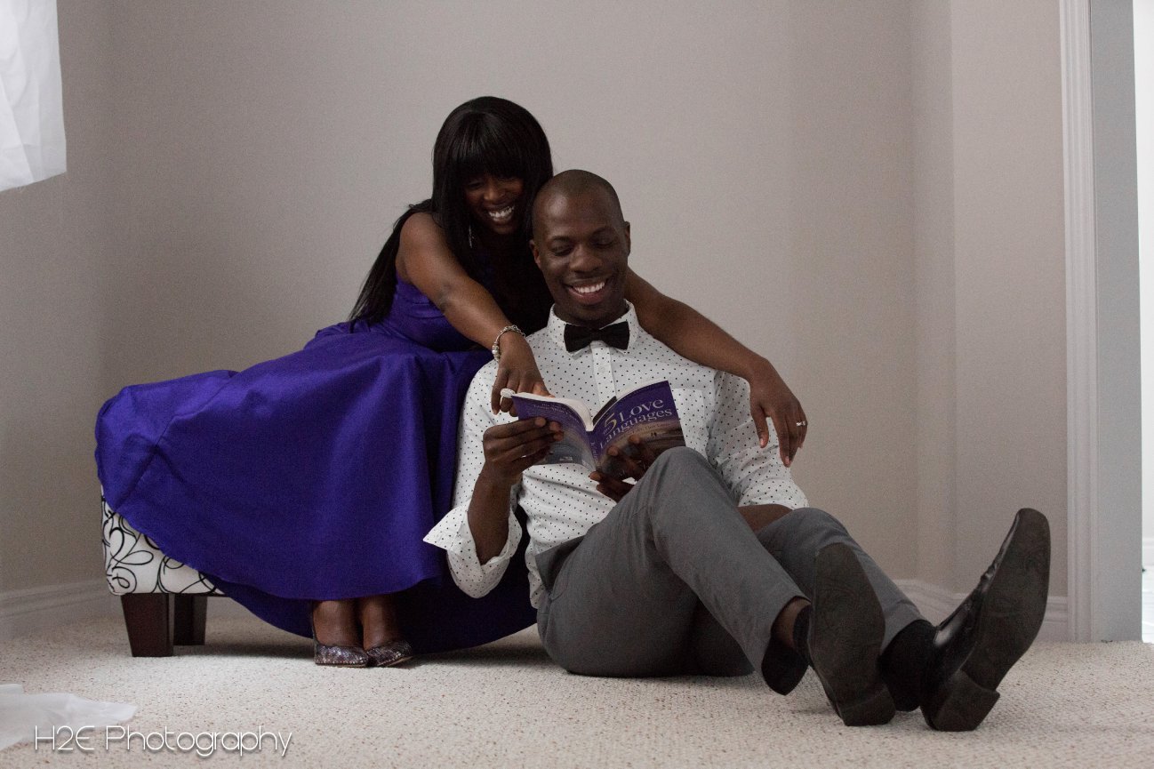 A couple sits on the floor, smiling while reading a book together. She's in a purple dress; he's in a bow tie and grey pants.