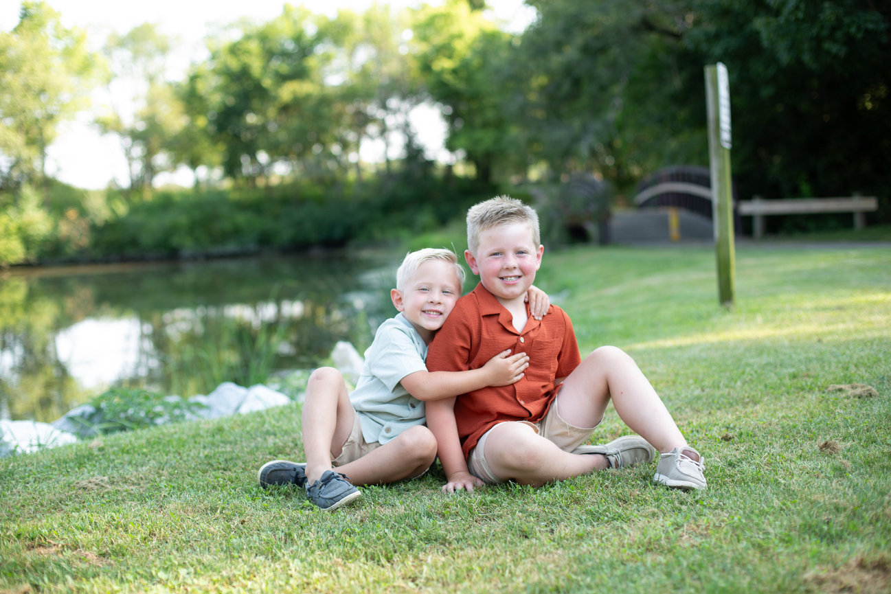 Two young boys sitting on the grass near a lake on a sunny day.