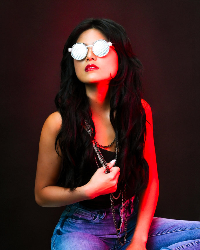 Person with long black hair wearing reflective sunglasses and layered necklaces, posed against a dark background with red lighting.