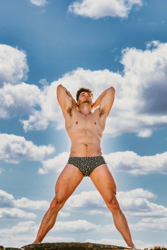 Muscular man posing with arms raised, wearing swim trunks, against a blue sky with clouds.