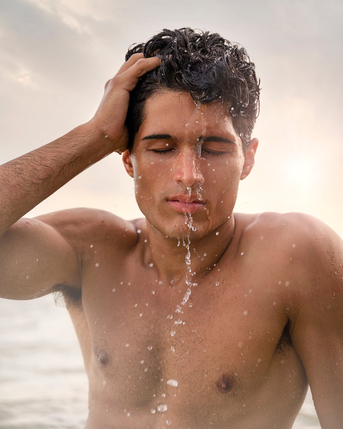 Young man with wet hair stands in water, water cascading down his face, against a soft, sunny background.