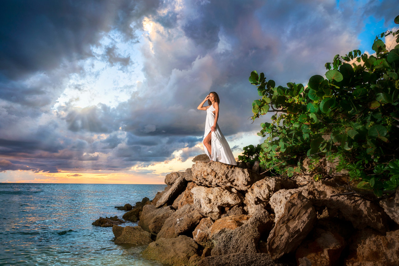 A woman in a flowing white dress stands on rocks by the ocean, with dramatic clouds and a sunset in the background.