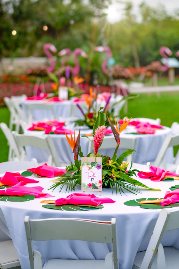 Decorative outdoor dining setup with floral centerpieces, pink napkins, and tropical decor for an event.