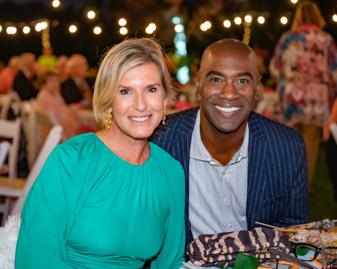 A smiling woman in a green dress and a man in a striped suit pose together at an outdoor event with festive lighting.