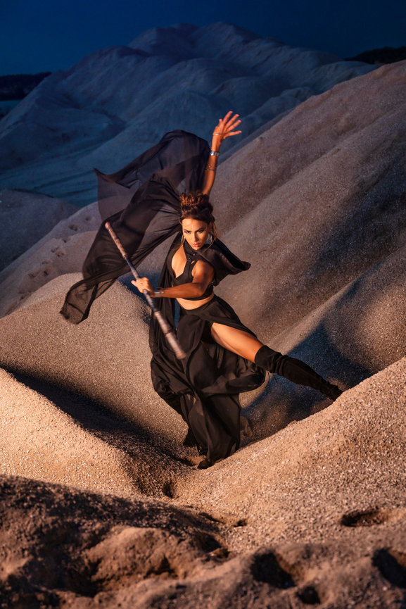 Dancer in a black outfit performing with a flowing fabric in a sandy landscape at dusk.