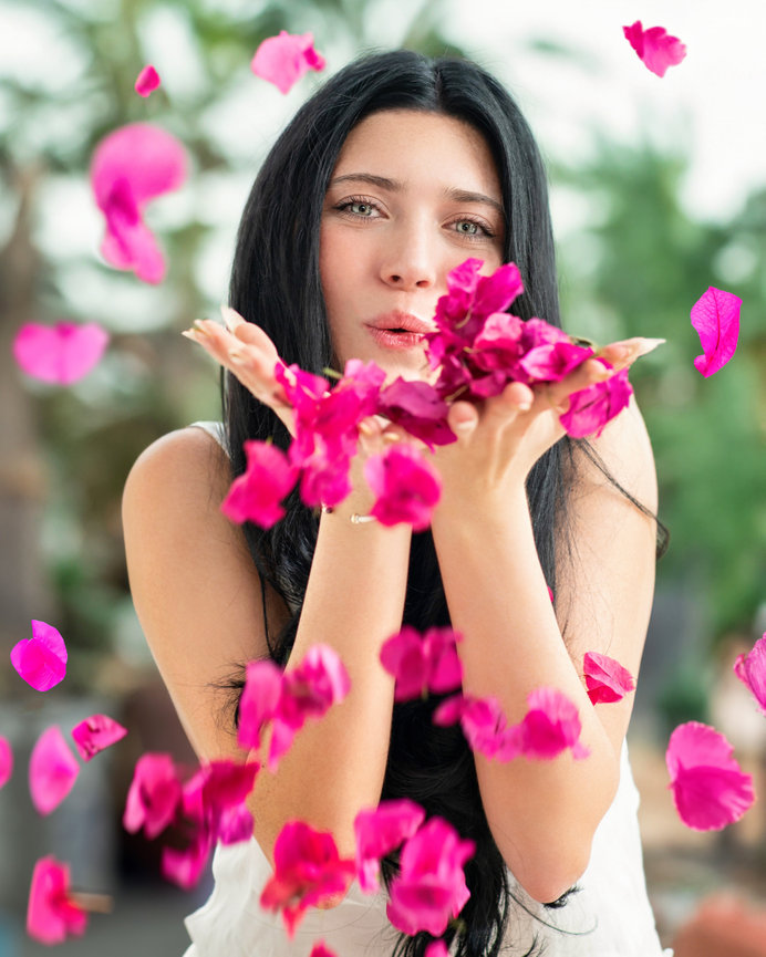 High school senior blowing pink flower petals into the air, with a lush green background.