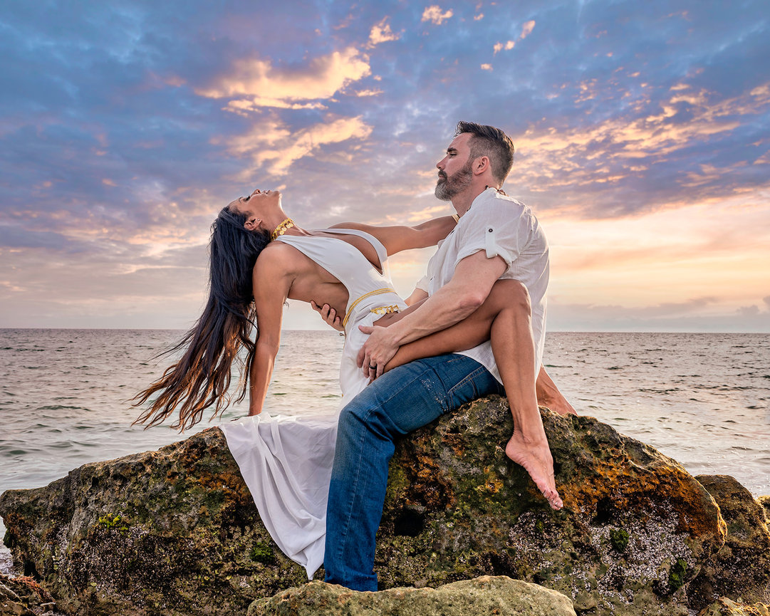 Couple in a romantic pose on a rocky shoreline at sunset, with ocean waves and colorful clouds in the background.