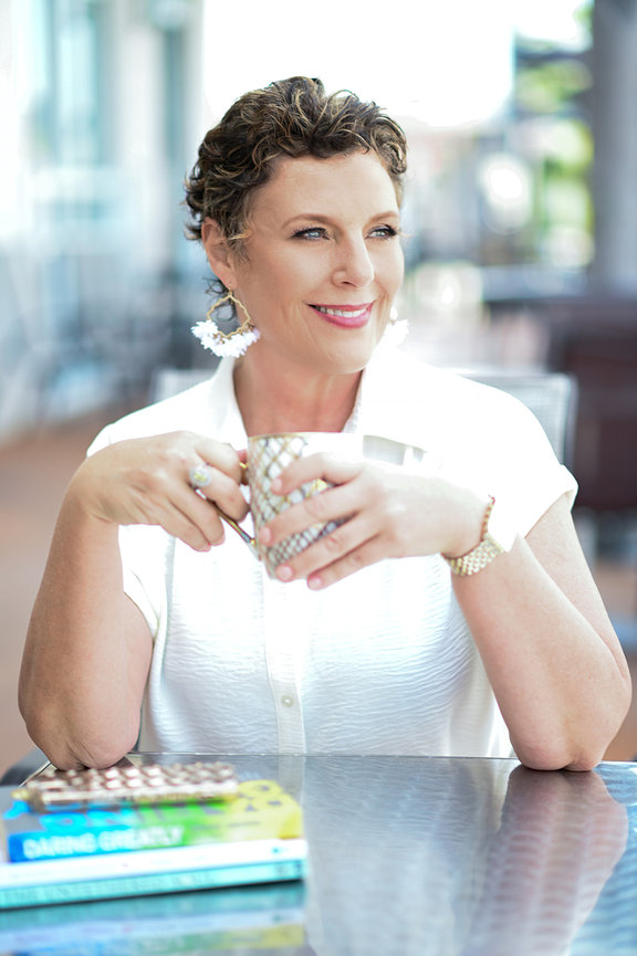 Woman with short curly hair, wearing a white shirt and large earrings, smiling while sitting at a table with books and a cup.