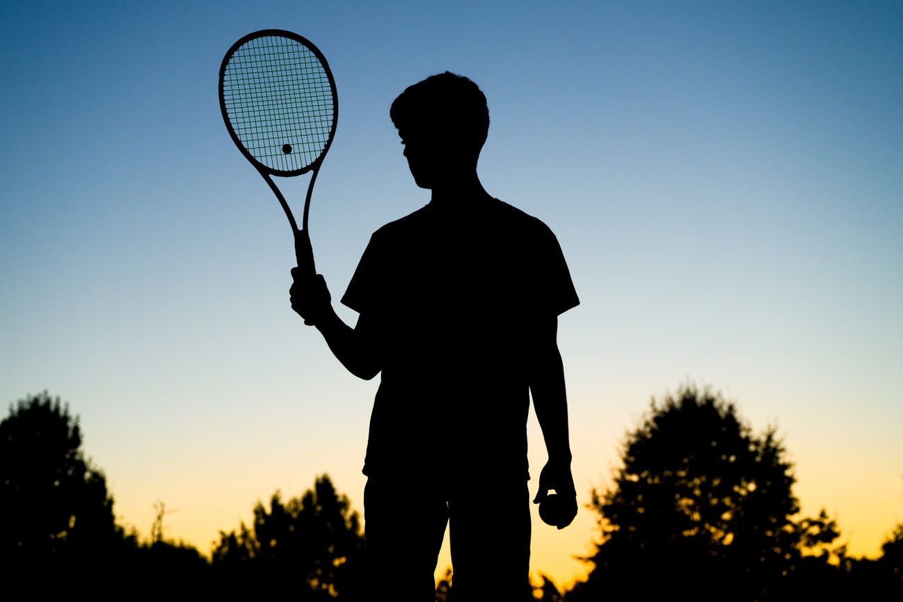 Silhouette of a senior holding a tennis racket and ball with a blue and yellow sunset behind