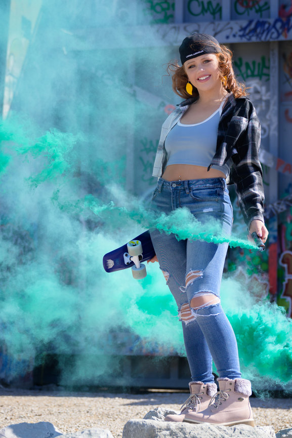 Senior wearing ripped jeans and crop top holding a skateboard with green smoke surrounding her
