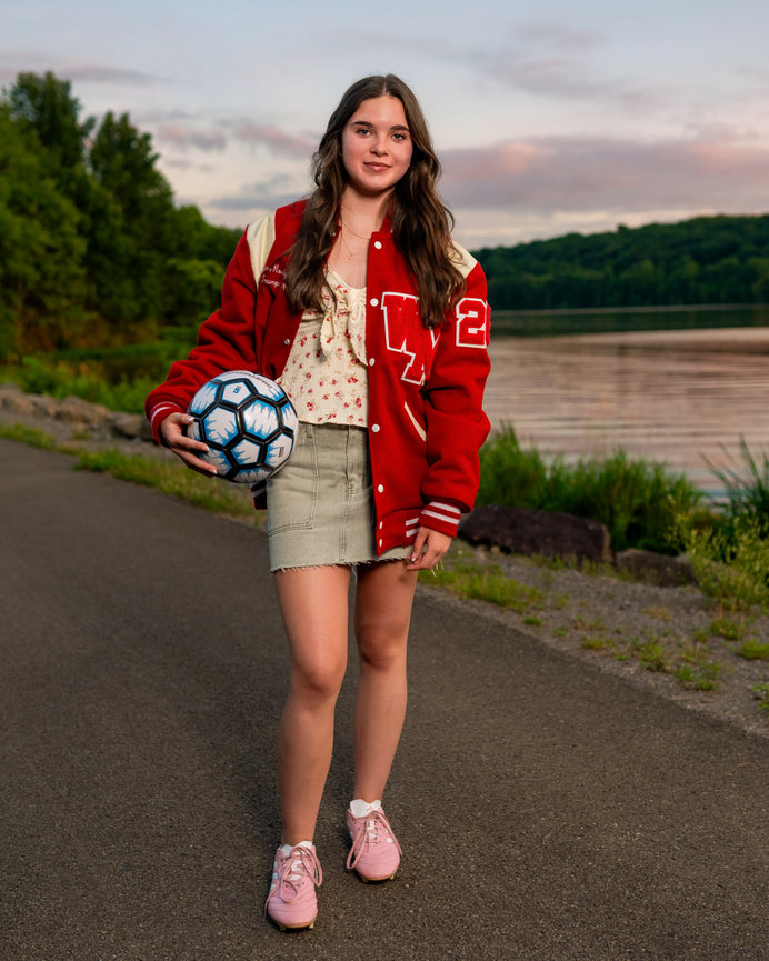 senior girl in her letterman jacket with her soccer shoes and ball in front of a lake