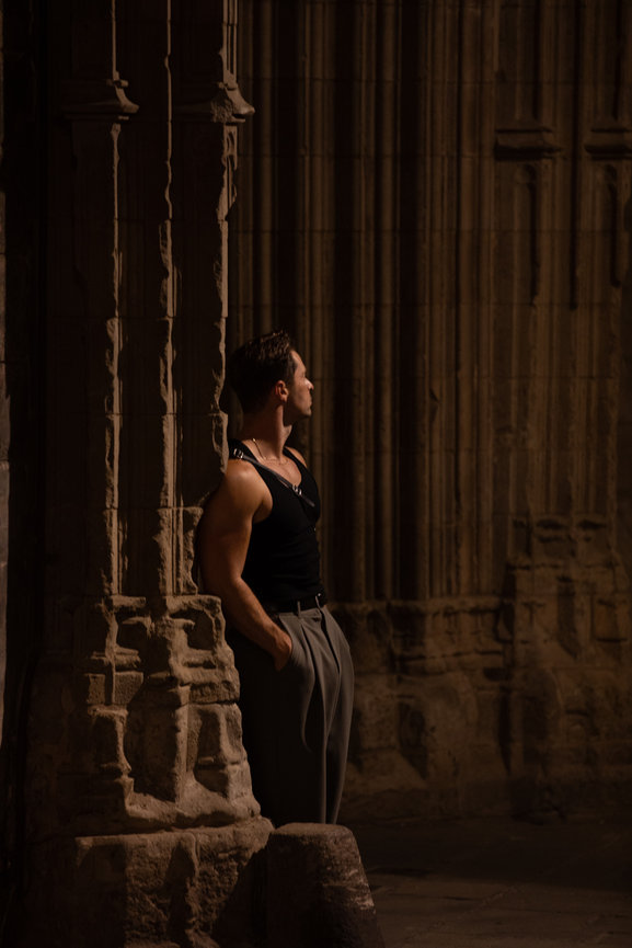 Man in a black tank top and gray pants leans against a stone pillar in a dimly lit, ornate architectural setting. Barcelona night