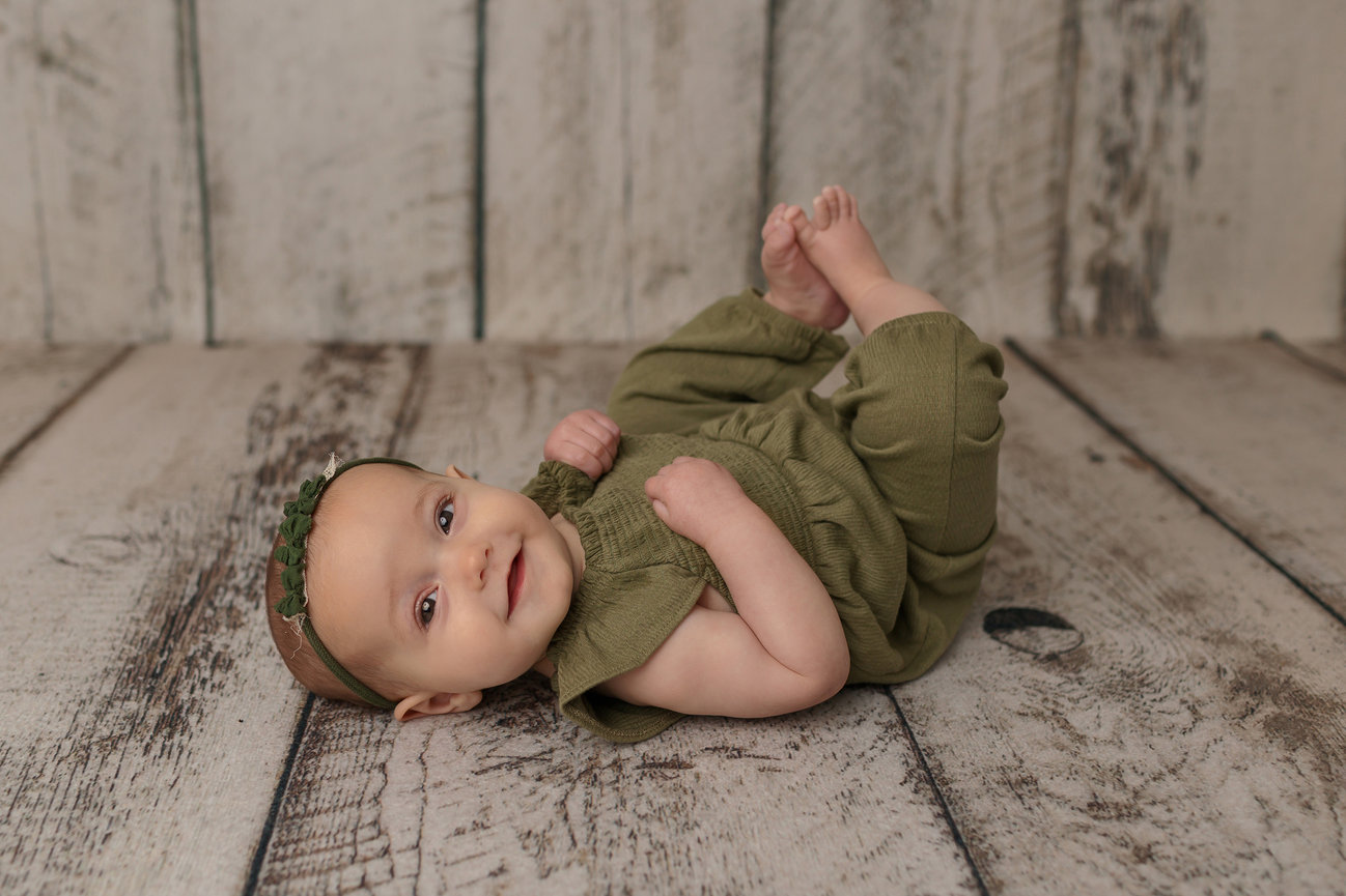 Baby in green outfit lying on wooden floor, smiling.