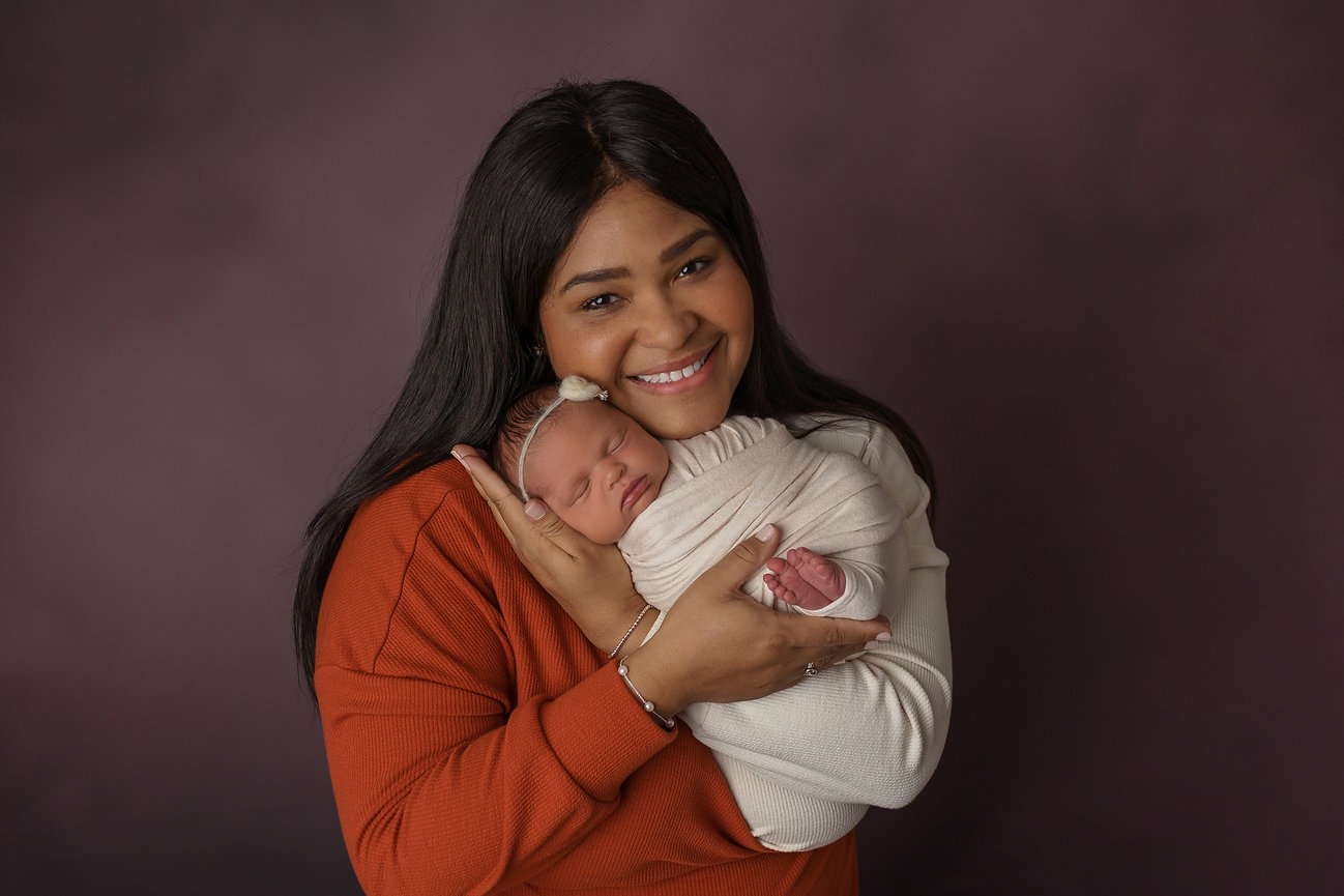 A woman smiling while holding a sleeping baby wrapped in a blanket against a dark background.