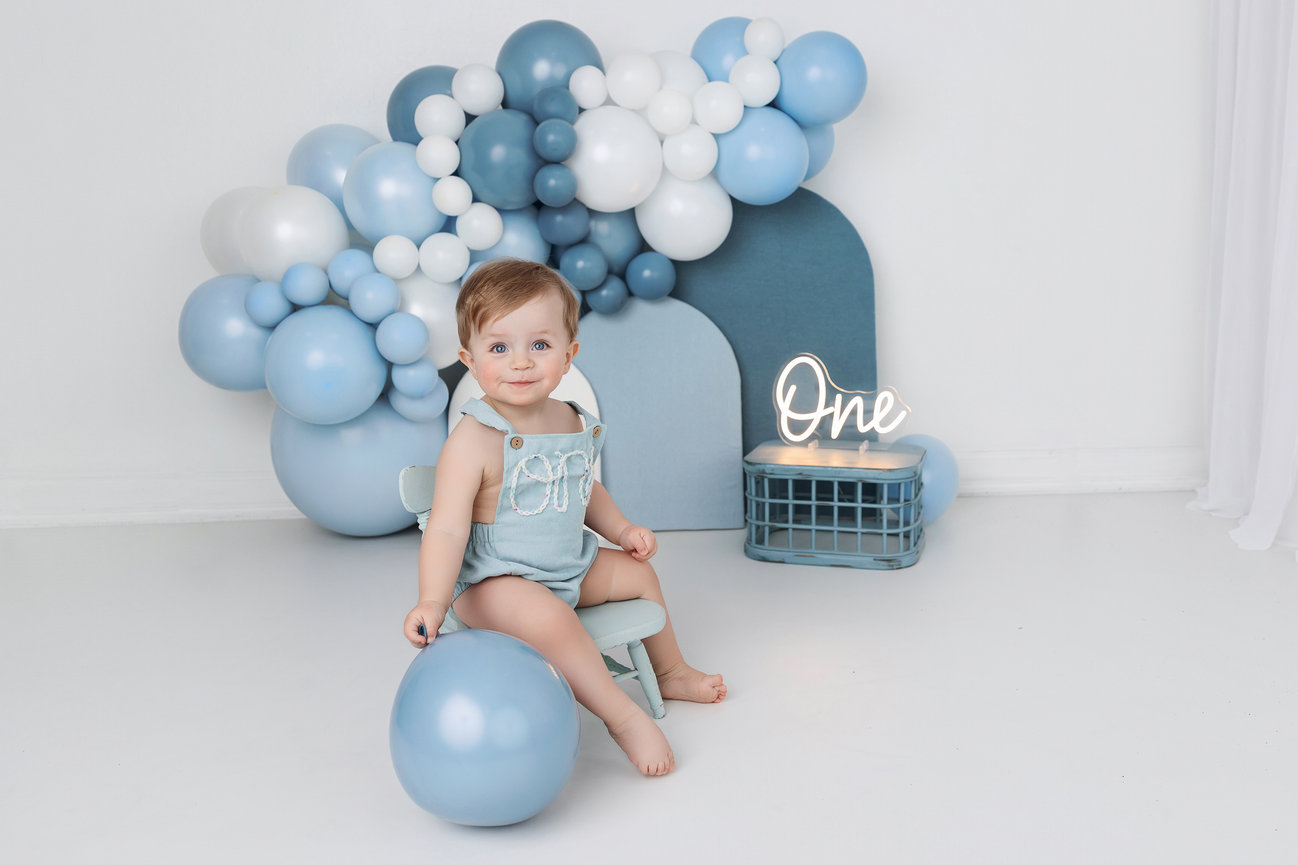 Baby sitting with blue balloons and One sign, celebrating first birthday in a light-colored room.
