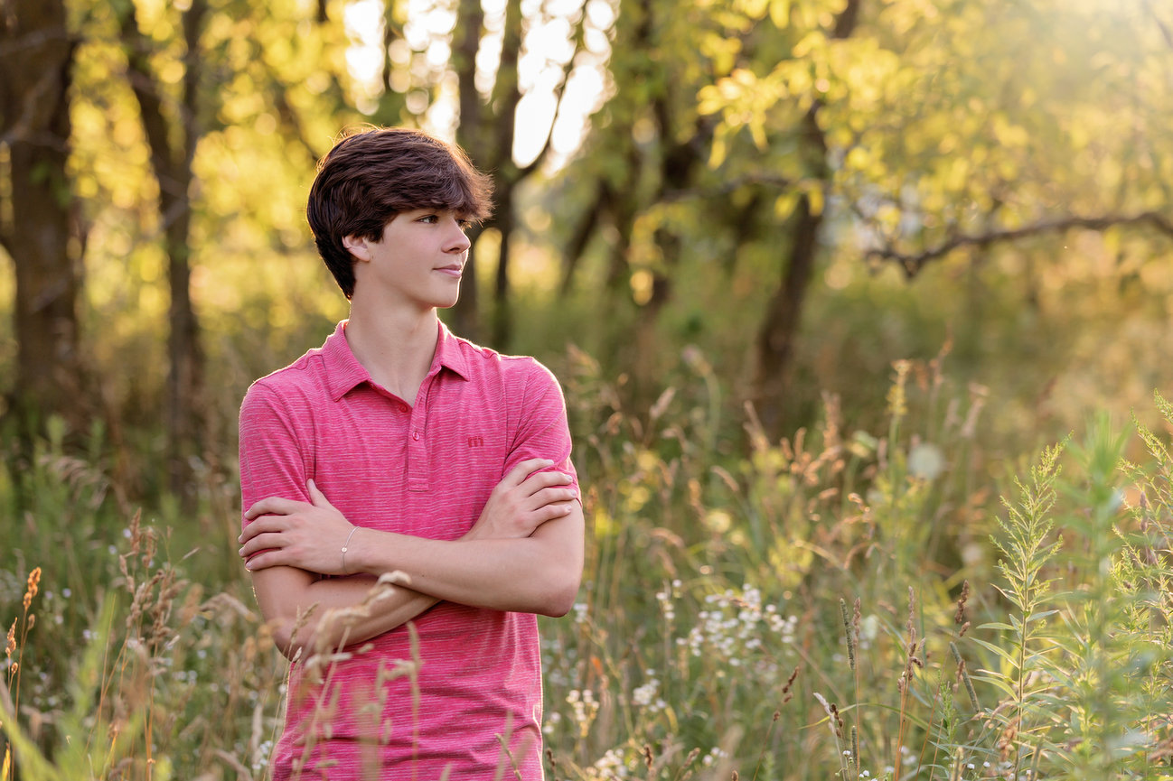 Young man in a pink shirt stands with arms crossed in a sunlit forest clearing.