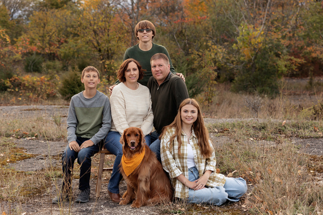 Family with a dog poses outdoors in autumn, surrounded by fall foliage.