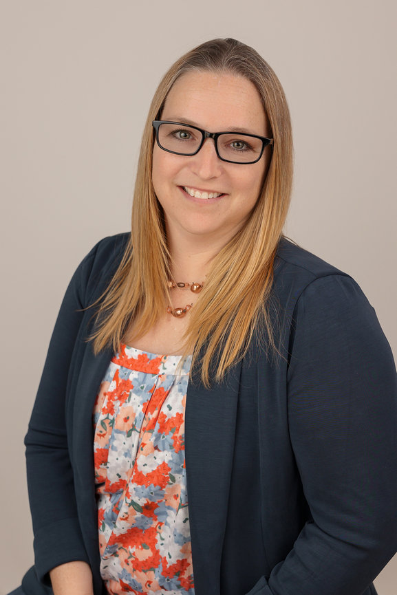 Woman with glasses smiling, wearing a floral blouse and navy blazer.