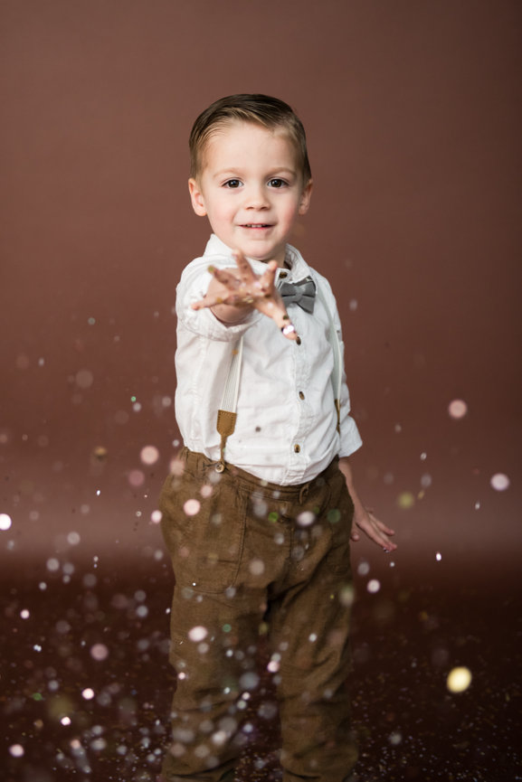 Young boy in formal attire with suspenders, smiling and reaching forward, surrounded by sparkling confetti against a brown backdrop for glitter mini session