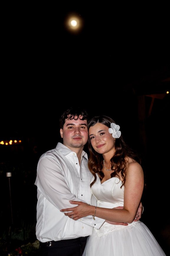 Young couple embracing at a nighttime wedding, dressed in white, with the woman wearing a flower hair accessory.
