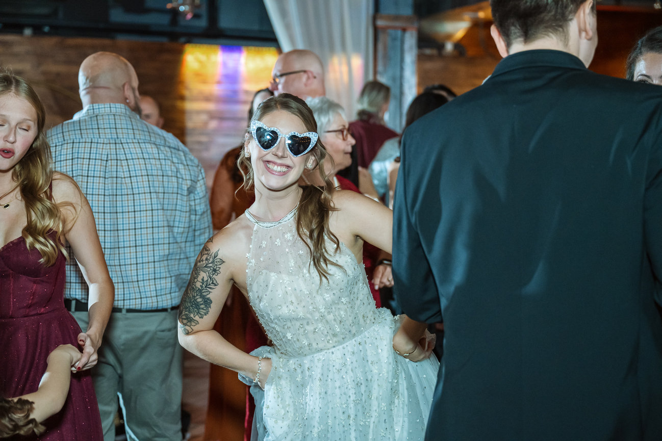 Smiling bride in heart-shaped sunglasses dancing at a wedding reception among guests