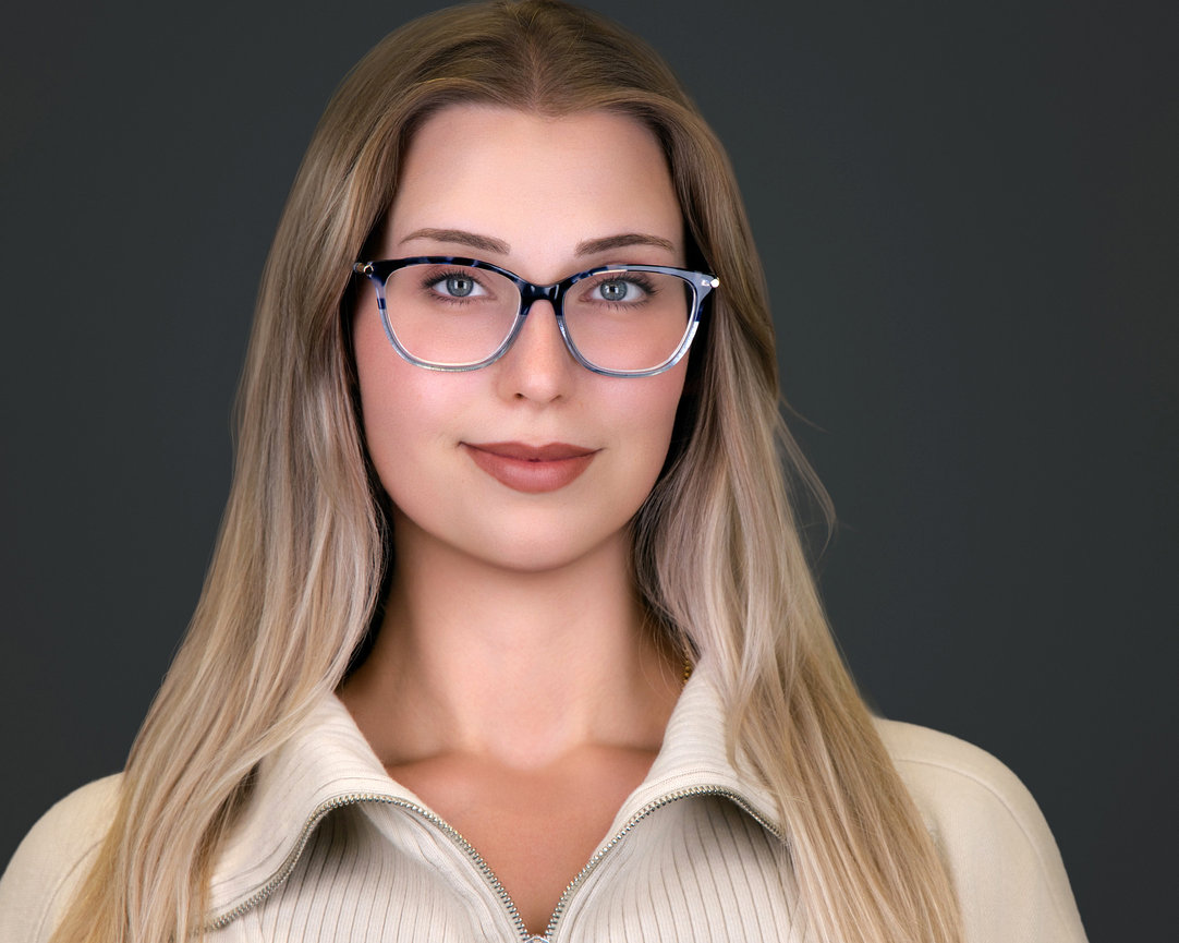 A woman with long blonde hair and glasses, wearing a beige zip-up sweater, against a gray background.