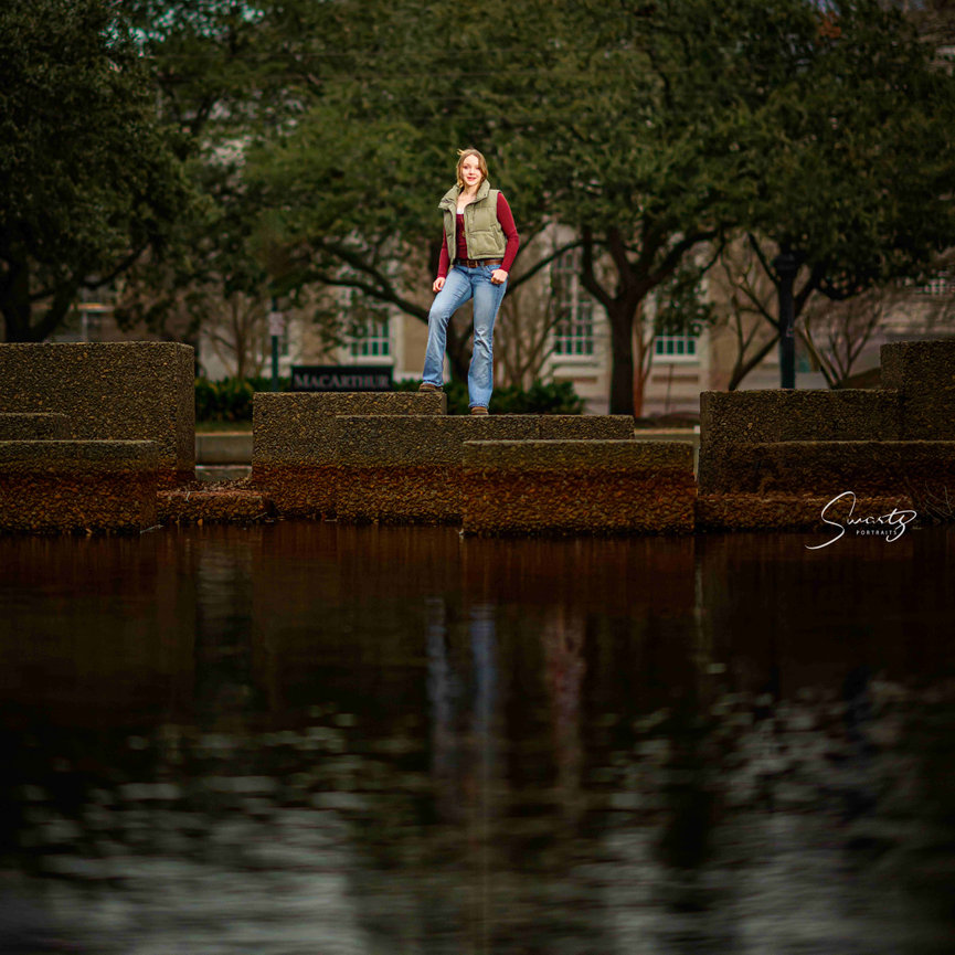 Senior girl on architectural concrete tiers reflected in MacArthur Park water with Texas live oaks framing — fine art environmental senior portrait by Master Photographer Ty Swartz, Swartz Portraits Katy Sugar Land Houston Texas