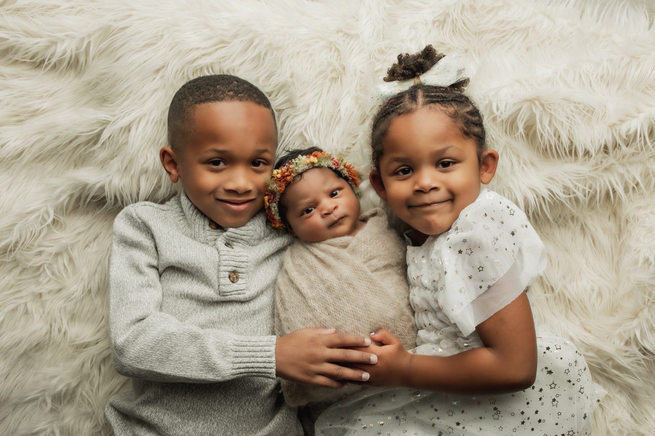 Two children holding a swaddled newborn on a fluffy blanket