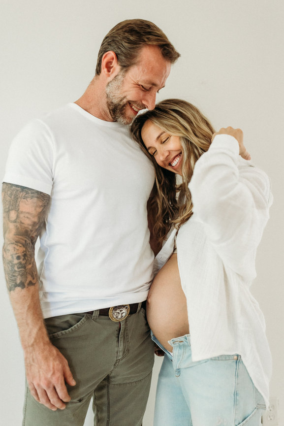 Expecting couple smiling; woman holds her belly, man with tattoos embraces her.