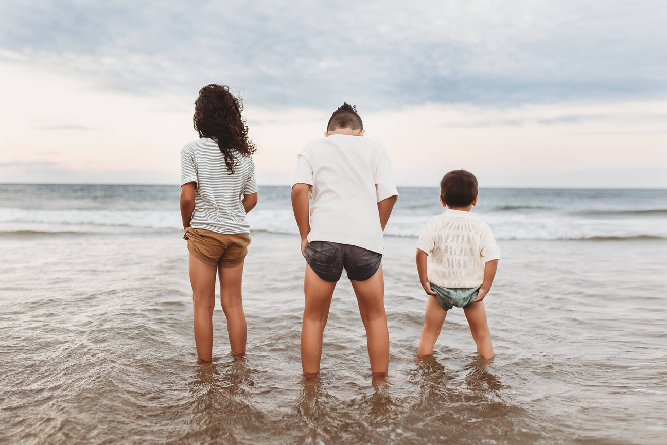 Three brothers standing ankle deep in the ocean during low tide in Sandy Hook, New Jersey. Captured by New Jersey Family Photographer Love of James Photography.
