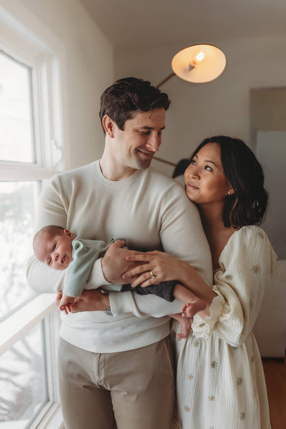 A father holds his infant while looking lovingly at his wife captured by Love of James Photography, New Jersey Newborn Photographer.