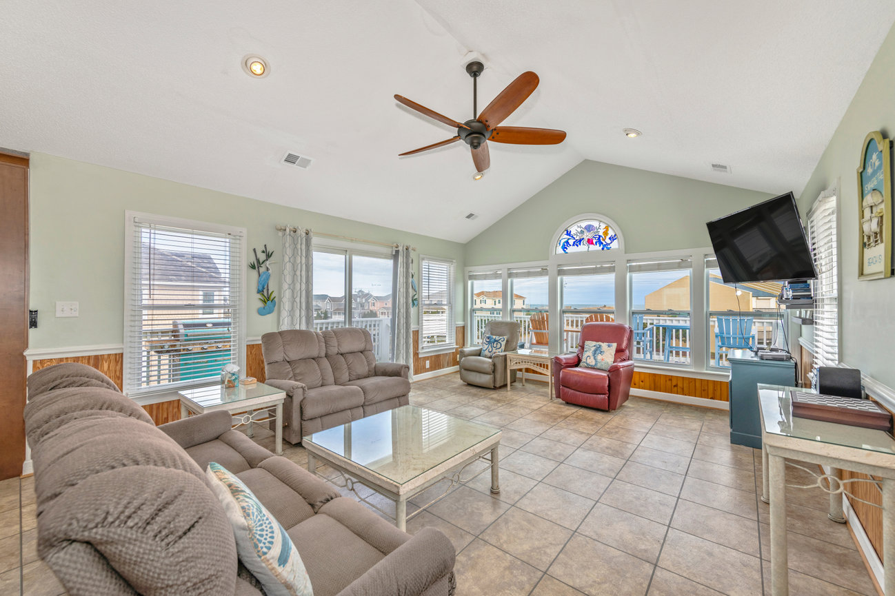 Spacious living room with large windows, comfy seating, ceiling fan, and TV, overlooking a coastal view.