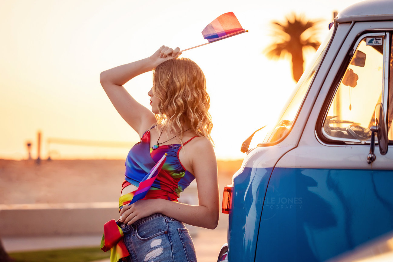 PROFILE OF A BLOND GIRL DRESSED IN RAINBOW COLORS HOLDING A PRIDE FLAG IN FRONT OF A VOLKSWAGEN BUS AT THE BEACH DURING GOLDEN HOUR SUNSET.