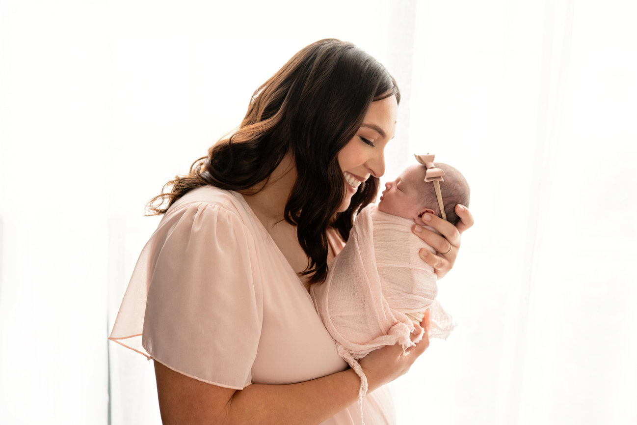 Smiling mother holding and kissing her swaddled newborn baby with a bow headband