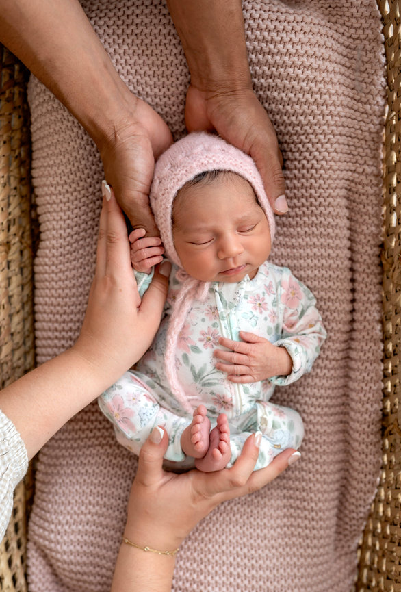 Newborn baby in floral outfit, gently held by adult hands on a pink knitted blanket.