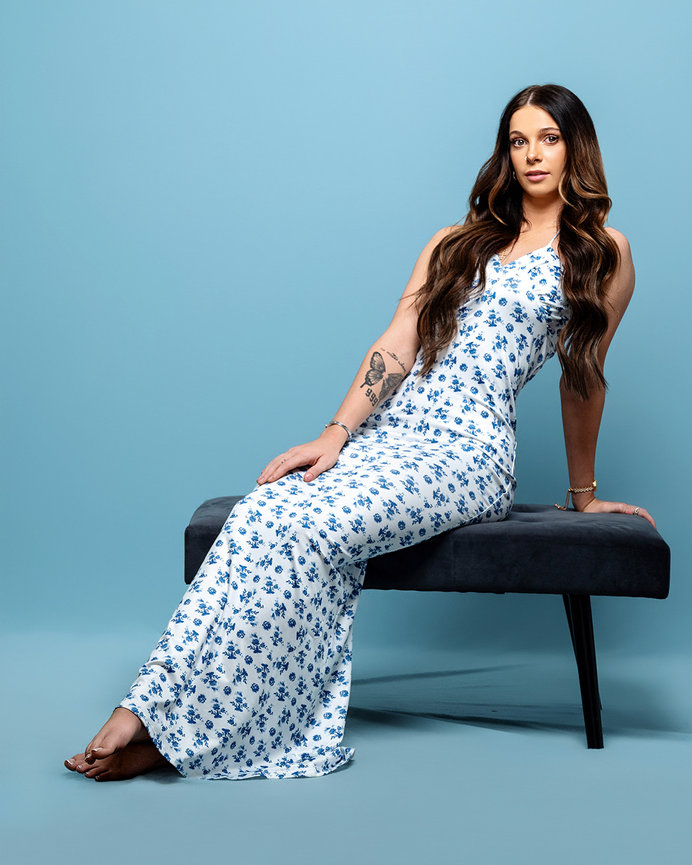 Model in a white dress with blue floral patterns sitting elegantly on a bench against a pastel blue backdrop in a Lehi, Utah studio. Captured by Tre Nelson Photo, showcasing high-class fashion and minimalist elegance.