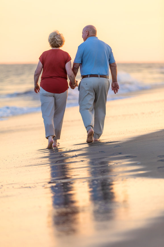 Grandpa and grandma walk down the beach holding hands in coordinated outfits at sunset with a water reflection.