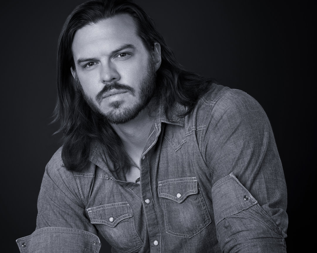 Black and white portrait of a man with long hair and a denim shirt, looking at the camera.