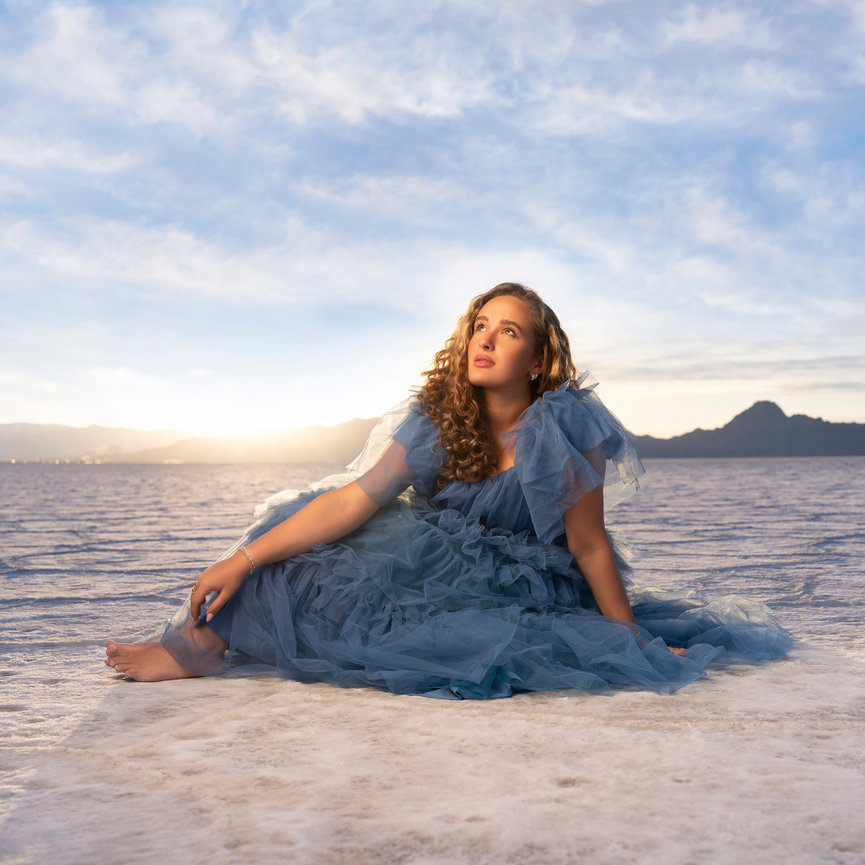 Senior girl in a blue dress sitting on salt flats at sunset with mountains in the background.