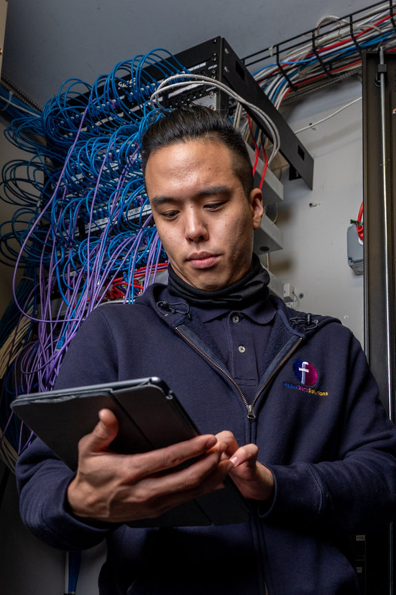 Technician using a tablet in a network server room with bundles of blue and purple cables.