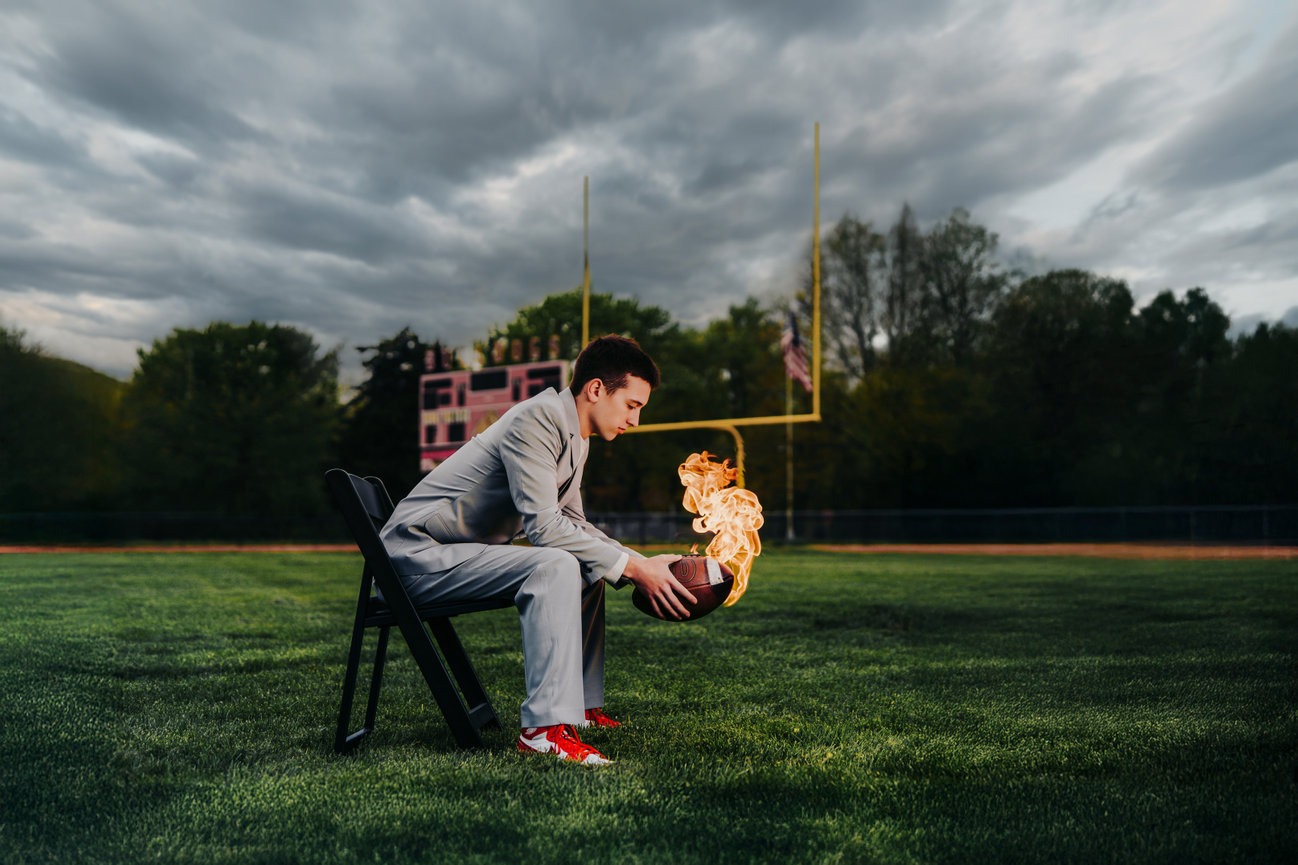 Young man in a suit sits on a football field holding a burning football under a cloudy sky