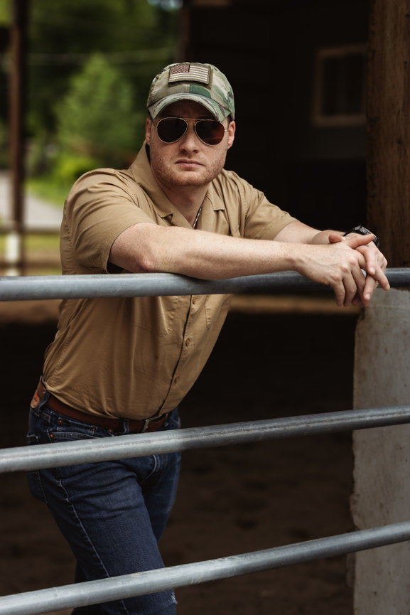 Man in sunglasses and camo cap leaning on a metal rail outdoors.