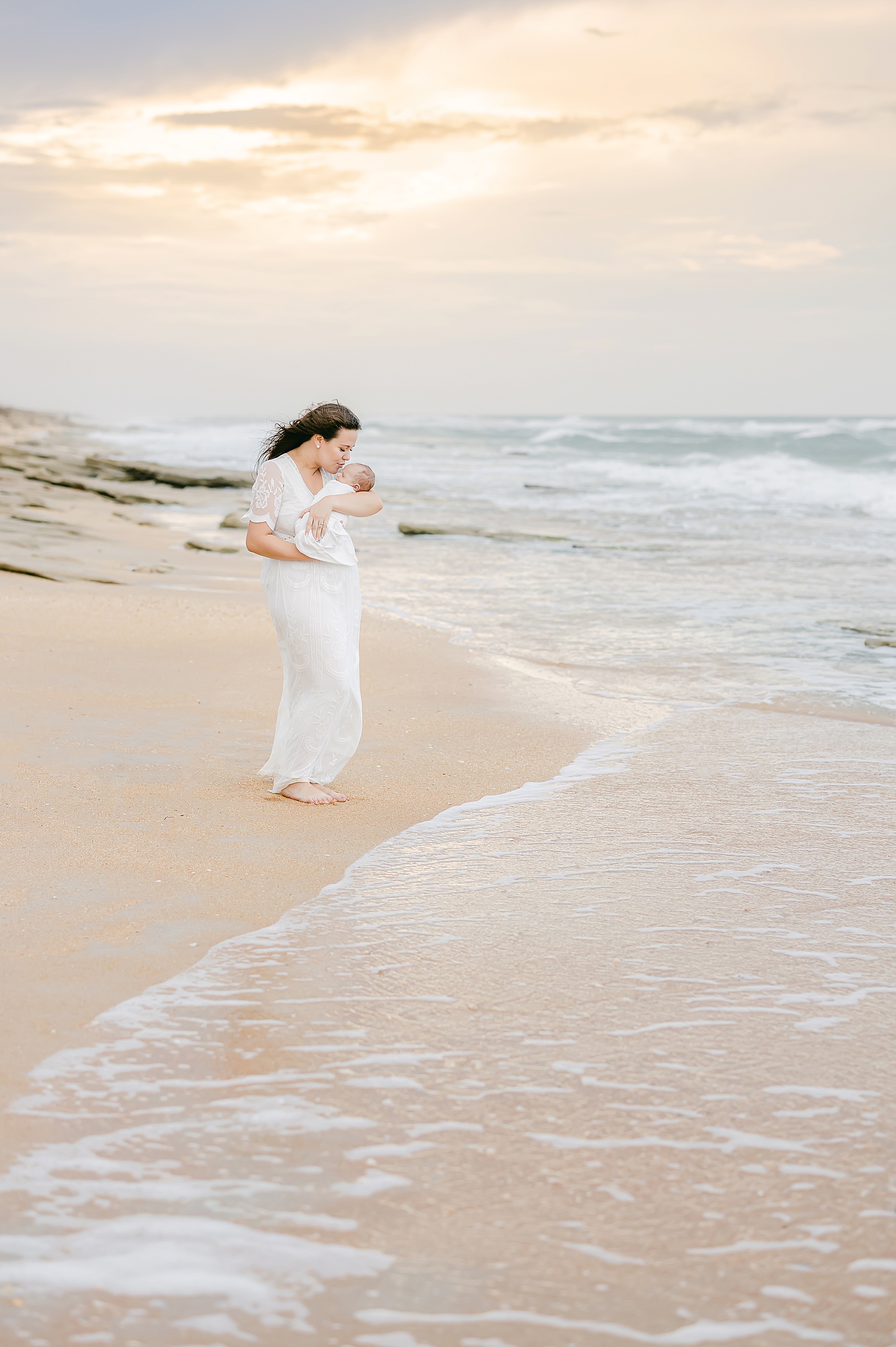 woman standing on beach in white long flowing dress in the sand as the surf comes in at sunset