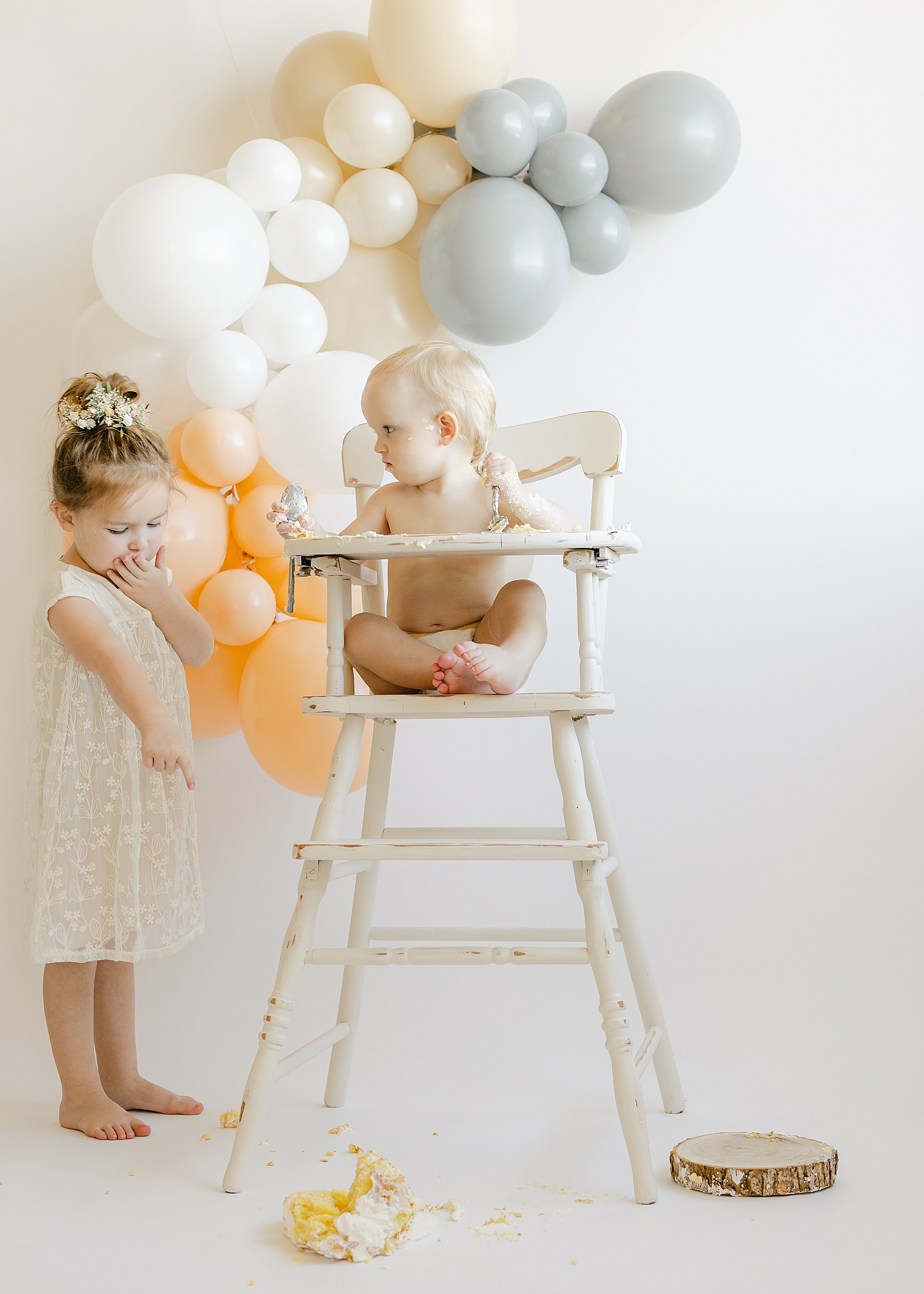 baby boy in white high chair throwing cake on ground with little girl looking and pointing in white dress