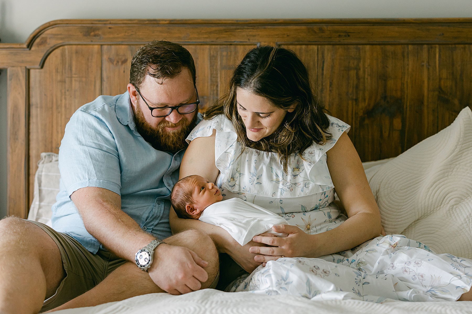 man and woman sitting on bed looking at newborn baby smiling