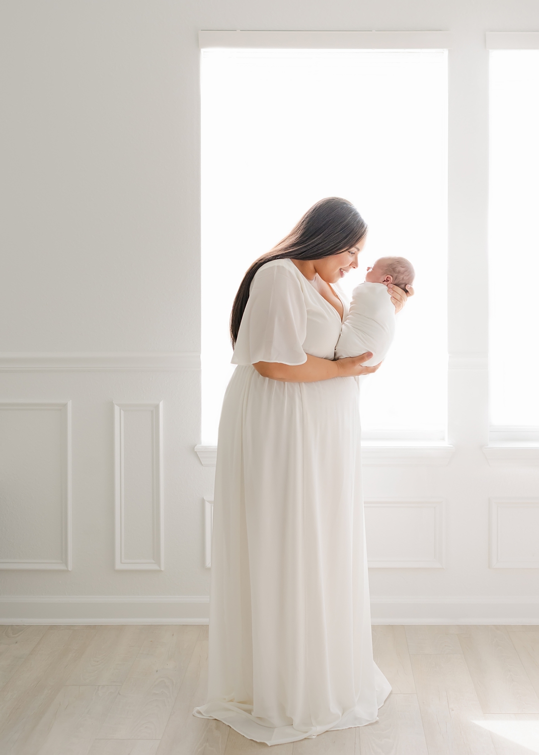 woman holding newborn baby in white blanket against backlit window