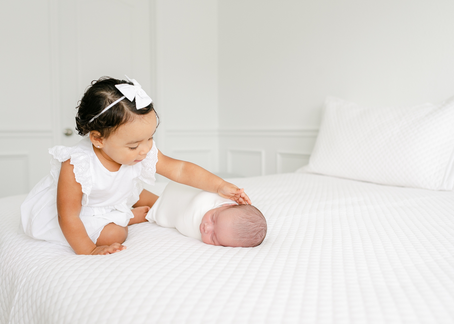 little girl dressed in white touching newborn baby boy on bed in white covers